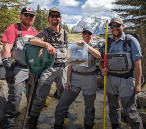 Four people wearing chest waders pose in a rocky stream with mountains in the background; one holds a green mesh net, another a bag of fish, and the others carry gear and a yellow querying pole or rod.