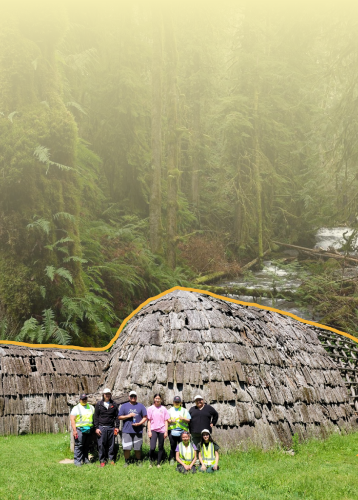 Group of people in safety vests posing in front of a large thatched longhouse in a misty forest clearing on green grass.
