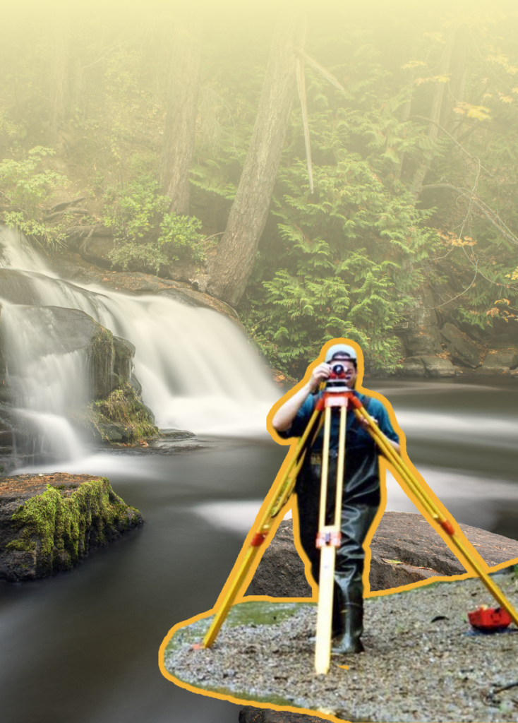Surveyor in blue shirt using a theodolite on a tripod by a waterfall along a rocky riverbank.