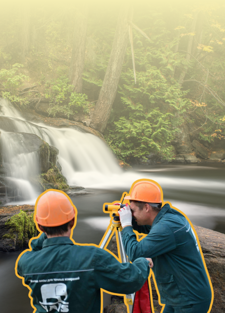 Two workers wearing orange hard hats survey a riverside site with a theodolite on a tripod, overlooking a forested waterfall.