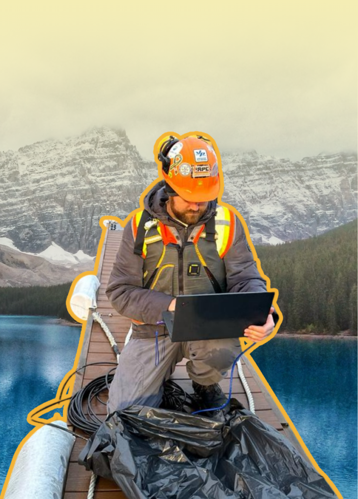 Field worker in safety gear kneeling on a bridge, using a laptop/tablet near a lake and snowy mountains in the distance, with an orange outline around him.