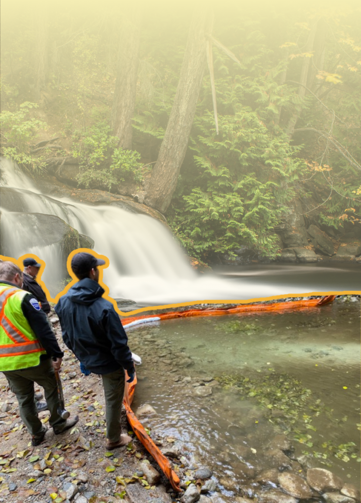 Workers in safety vests and jackets stand on a rocky riverbank, laying an orange containment boom by a small waterfall.
