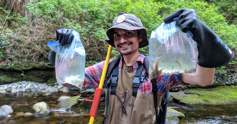 Smiling man in plaid shirt and hat holds two plastic bags with water samples near a rocky stream, wearing gloves and waders in a forested area.